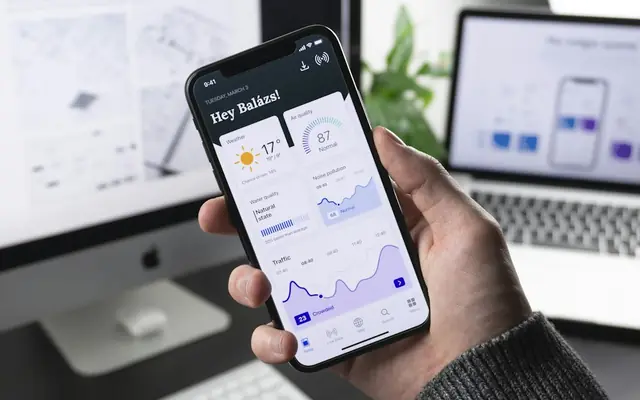 Hand holding a phone with a mobile app open, laptop in the background